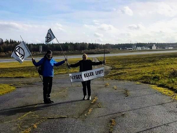 Two climate activists standing on an airport runway, holding a banner und flags.